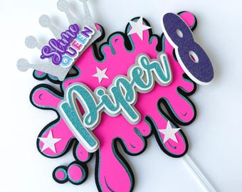 May include: A pink and black cake topper with the name "Piper" in teal and silver glitter. The topper includes a silver crown with the words "Slime Queen" and the number "8". White stars are scattered across the pink splash design.