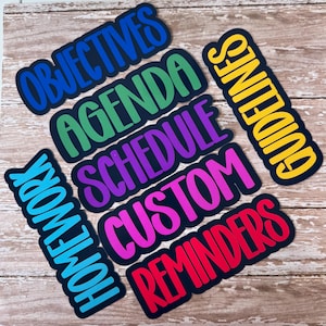 May include: A collection of colorful, die-cut word labels on a wooden surface. The labels include words like "Objectives," "Agenda," "Schedule," "Custom," "Reminders," "Homework," and "Guidelines," each with a unique color scheme and a black border.