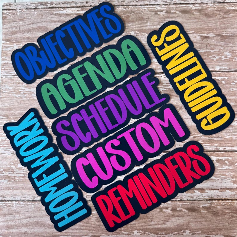 May include: A collection of colorful, die-cut word labels on a wooden surface. The labels include words like "Objectives," "Agenda," "Schedule," "Custom," "Reminders," "Homework," and "Guidelines," each with a unique color scheme and a black border.