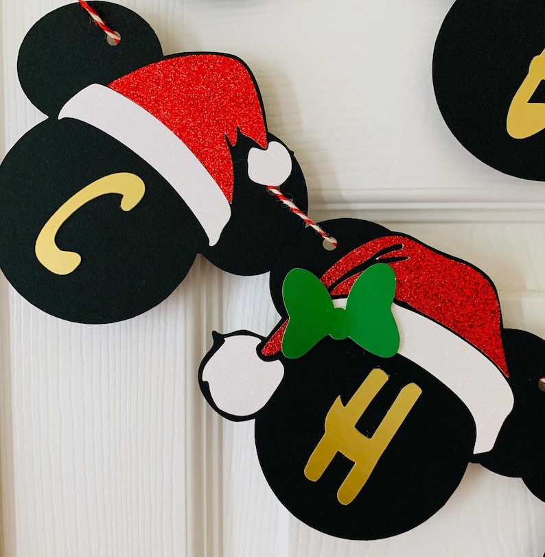 以下が含まれることがあります： 黒と金の文字で「クリスマス」と綴られたクリスマスバナー。各文字は別々の黒い円形に書かれており、各円形には赤と白のサンタ帽と白いポンポンが付いています。円形は赤と白の紐で繋がっています。