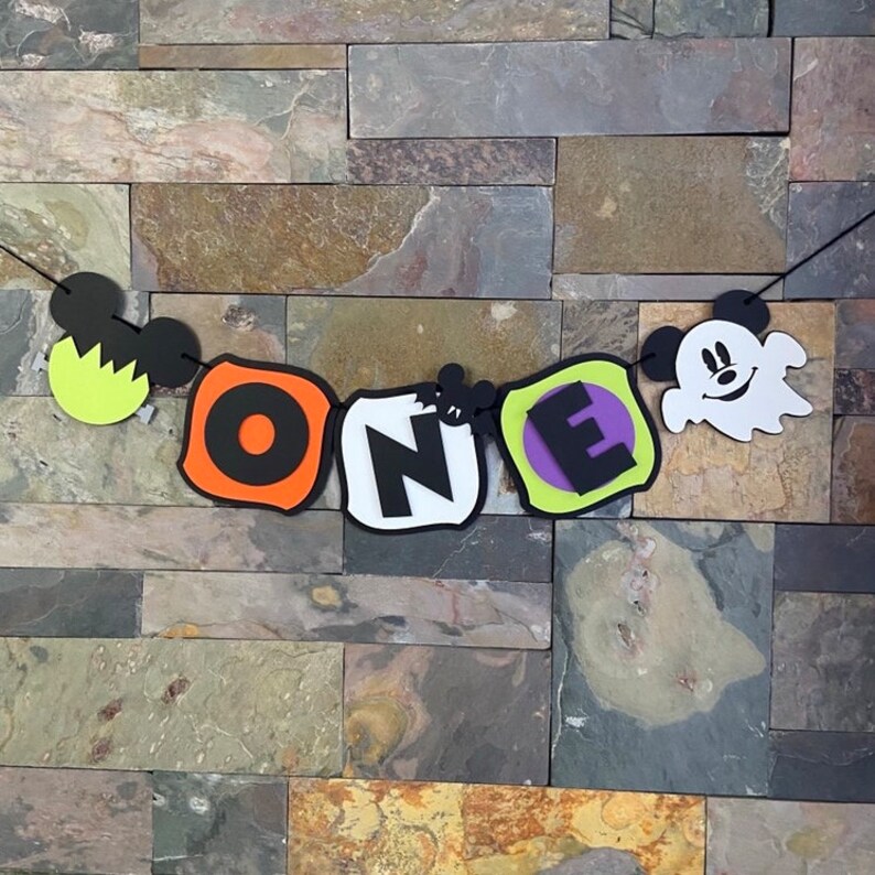 Puede incluir: Una pancarta de Halloween con la palabra "ONE" en letras negras sobre fondos coloridos. La pancarta presenta personajes de Mickey Mouse y fantasmas.