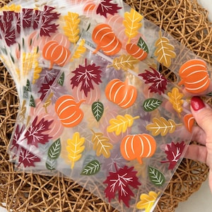May include: A stack of clear plastic zip-lock bags featuring a repeating autumn-themed pattern of orange pumpkins, deep red maple leaves, yellow oak leaves, and dark green foliage. The bags are transparent with a smooth, flexible texture.