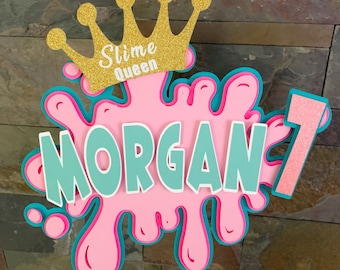 May include: A pink and turquoise slime-themed birthday party decoration with the name "MORGAN" and the number "1" in glitter. The decoration also features a gold crown with the words "Slime Queen" on it.