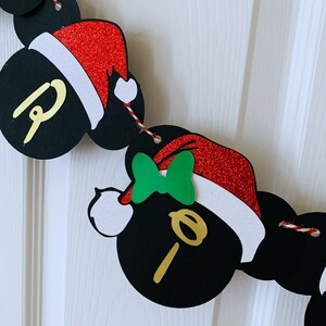以下が含まれることがあります： ミッキーマウスの耳とサンタ帽子のついた黒と赤のクリスマスガーランド。ガーランドは金色のグリッターのアクセントと緑のリボンが付いています。