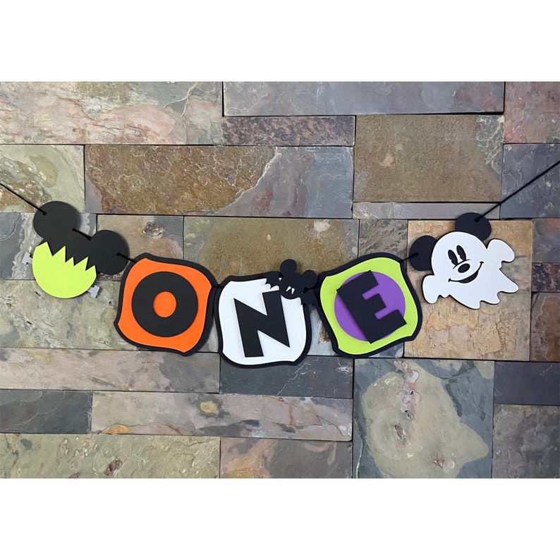 Puede incluir: Bandera de tem&aacute;tica de Halloween con la palabra "ONE" en letras negras sobre cuadrados blancos, con acentos naranja, verde y morado. La bandera tambi&eacute;n presenta una cabeza de Mickey Mouse negra con un fantasma amarillo y un fantasma blanco con una cabeza de Mickey Mouse negra.