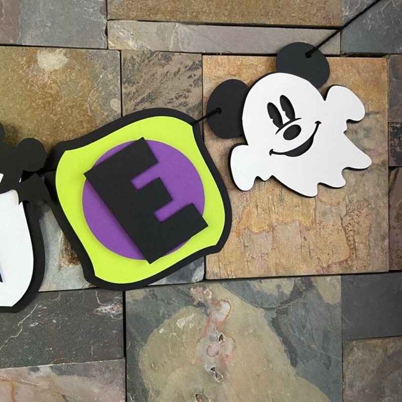 Puede incluir: Un banner de Halloween con una letra "E" verde, morada y negra y un fantasma blanco con orejas negras de Mickey Mouse.
