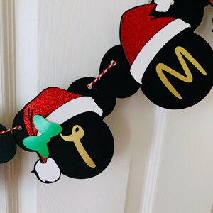 以下が含まれることがあります： ミッキーマウスの耳と赤いサンタ帽子のついた黒と金のクリスマスバナー。バナーには金色の文字で「メリー」と書かれています。