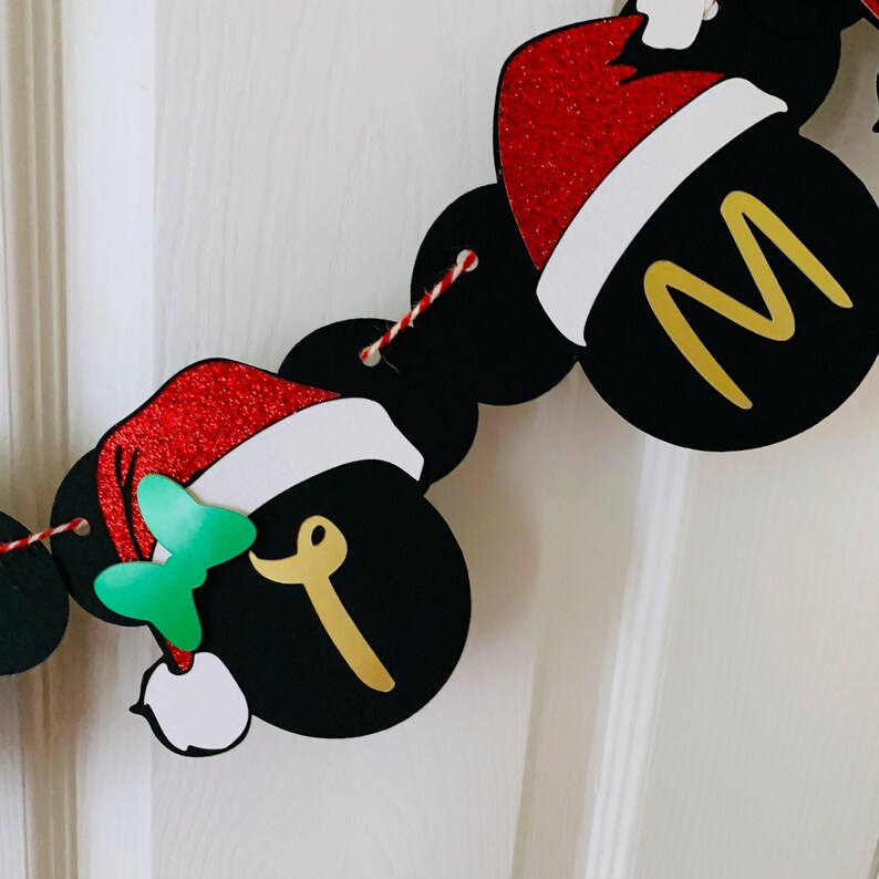 以下が含まれることがあります： ミッキーマウスの耳と赤いサンタ帽子のついた黒と金のクリスマスバナー。バナーには金色の文字で「メリー」と書かれています。