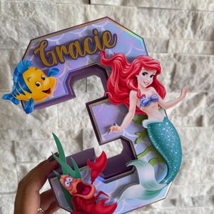 May include: A custom-made number 3 cake topper featuring Ariel the mermaid, Flounder, and Sebastian from The Little Mermaid. The number is purple with gold accents and the name "Gracie" in gold script.