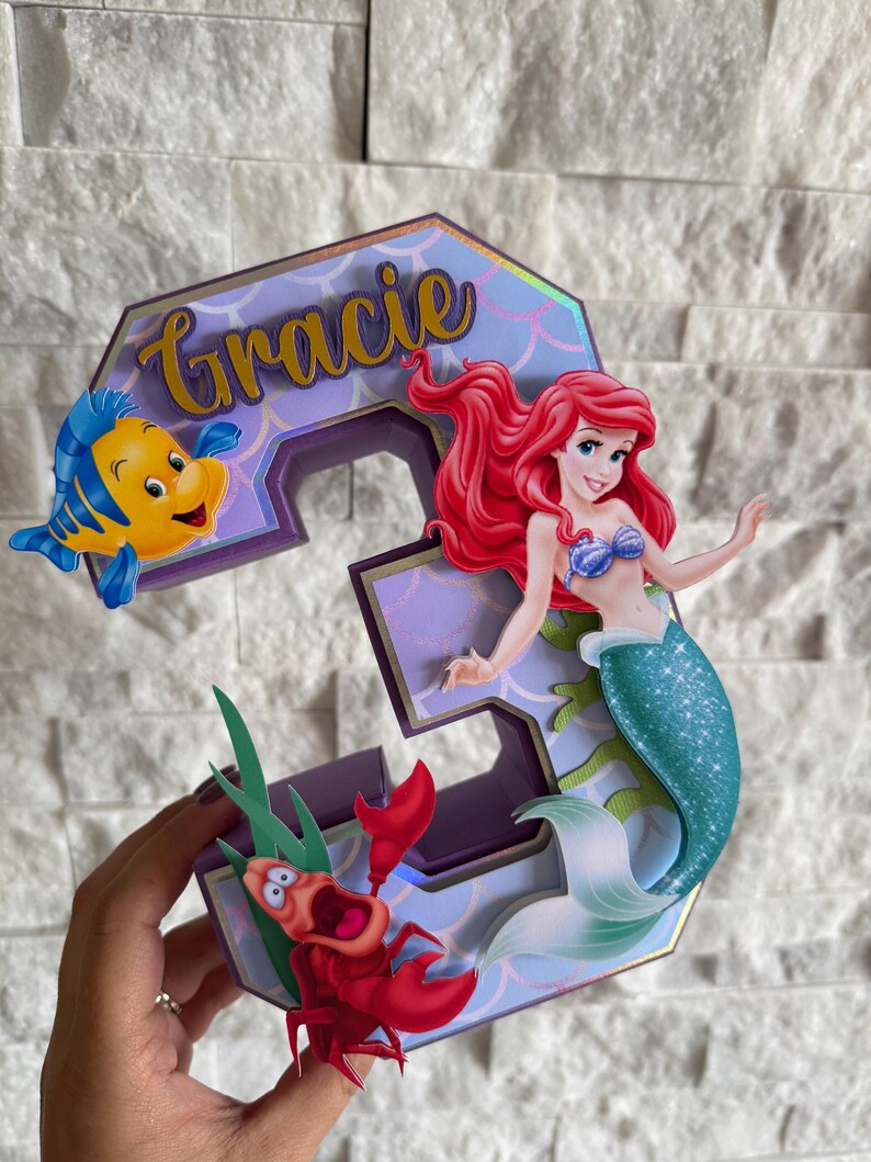 May include: A custom-made number 3 cake topper featuring Ariel the mermaid, Flounder, and Sebastian from The Little Mermaid. The number is purple with gold accents and the name "Gracie" in gold script.