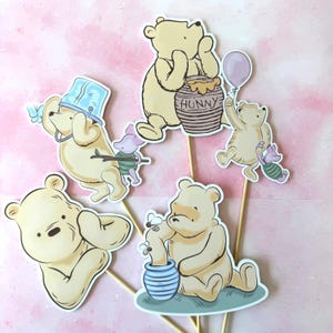 Pode incluir: Decorações de festa com o tema Ursinho Pooh. As decorações apresentam ilustrações do Ursinho Pooh, Leitão e potes de mel. Os personagens estão em várias poses, alguns segurando balões e potes de mel. As decorações estão em palitos de madeira.