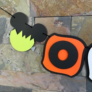 Puede incluir: Bandera de Halloween con orejas de Mickey Mouse en blanco y negro, un monstruo de Frankenstein verde, un c&iacute;rculo naranja y negro y un fantasma blanco.
