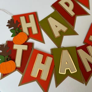May include: A Happy Thanksgiving banner with brown, red, and green pennants. Each pennant features a large, cream-colored letter with gold accents. The letters spell out "Happy Thanksgiving". The banner is decorated with small, orange pumpkins and brown leaves.