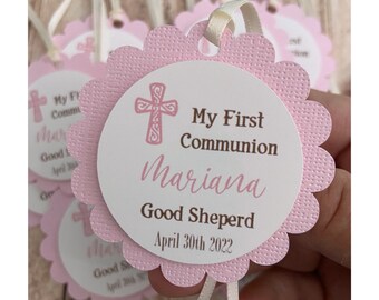 May include: Pink and white scalloped circle favour tag with a brown cross, the text "My First Communion", the name "Mariana", and the text "Good Sheperd April 30th 2022".
