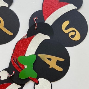 以下が含まれることがあります： 黒いミッキーマウスの頭の切り抜きがついたクリスマスバナー。それぞれに赤と白のサンタの帽子、金の文字、緑のリボンが付いています。文字は「SANTA」と綴られています。