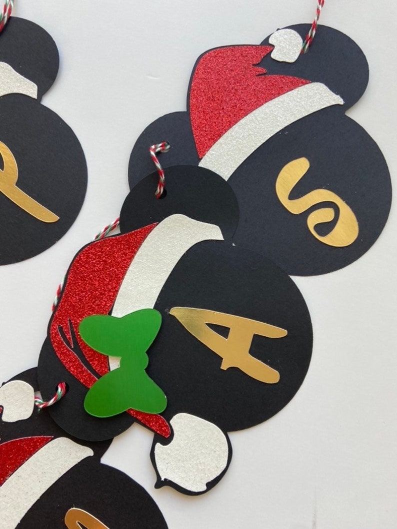 以下が含まれることがあります： 黒いミッキーマウスの頭の切り抜きがついたクリスマスバナー。それぞれに赤と白のサンタの帽子、金の文字、緑のリボンが付いています。文字は「SANTA」と綴られています。
