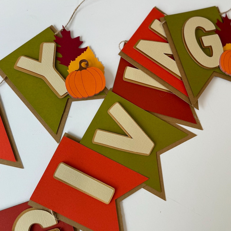 May include: A Thanksgiving banner with the word "Giving" spelled out in brown, red, and green felt letters. Each letter is attached to a brown triangle with a green felt backing. A small orange felt pumpkin with a green stem is attached to the "G" letter.