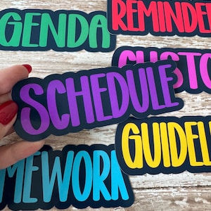 May include: A collection of colorful word stickers with black borders. The stickers feature words like "OBJECTIVES," "AGENDA," "REMINDER," and "SCHEDULE" in various bright colors. The stickers are held by a hand with red nail polish.