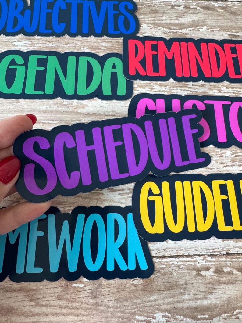 May include: A collection of colorful word stickers with black borders. The stickers feature words like "OBJECTIVES," "AGENDA," "REMINDER," and "SCHEDULE" in various bright colors. The stickers are held by a hand with red nail polish.