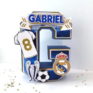 May include: A blue and white letter "G" cake topper with the name "GABRIEL" in blue and gold. Includes a soccer jersey with the number 8, a soccer ball, a trophy, and a team logo.