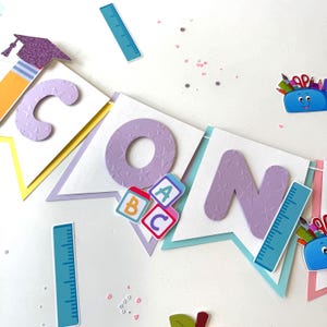 Puede incluir: Una pancarta morada y blanca con la palabra "SCHOOL" en letras moradas grandes. La pancarta está decorada con útiles escolares coloridos, incluyendo un lápiz, una regla, una mochila y bloques de alfabeto.