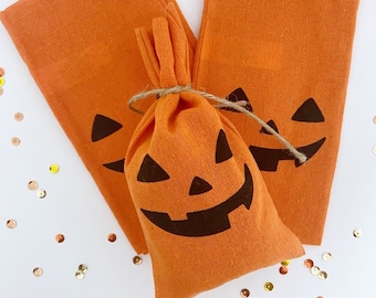 May include: Orange Halloween-themed fabric items. The image shows two orange napkins and a small drawstring bag, all featuring a black jack-o'-lantern face design. The bag is tied with twine. Gold confetti is scattered around the items.