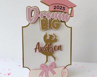 May include: A pink and gold graduation cake topper with the text "Dream Big" and the year "2025". The topper also features a silhouette of a graduate with the name "Andrea" and a graduation cap and diploma.