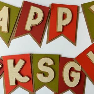 May include: A banner with the words "Happy Thanksgiving" in white letters with a wood grain texture on a red and green background. The letters are attached to brown paper pennants.