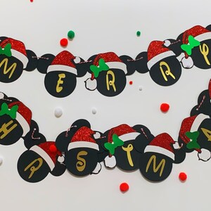 以下が含まれることがあります： 赤と白のサンタ帽と、金色の文字で「メリークリスマス」と綴られた黒いミッキーマウスの耳が付いたクリスマスバナーです。