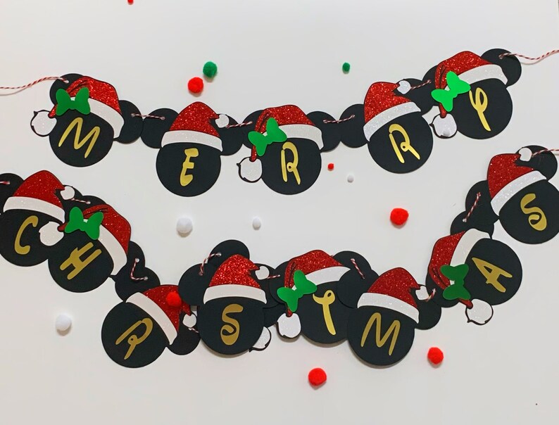以下が含まれることがあります： 赤と白のサンタ帽と、金色の文字で「メリークリスマス」と綴られた黒いミッキーマウスの耳が付いたクリスマスバナーです。