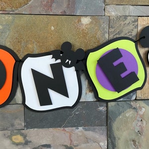 Puede incluir: Bandera de Halloween con la palabra "ONE" en letras negras sobre fondos blanco y verde. Un murci&eacute;lago negro con colmillos cuelga de la letra "N".