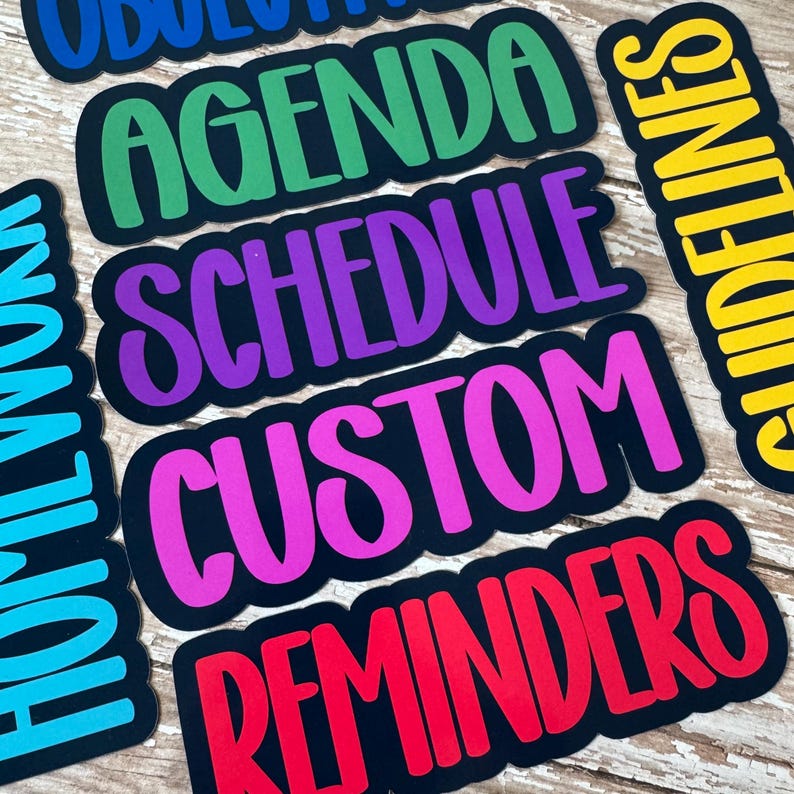 May include: A collection of colorful planner stickers with various words, including "Agenda," "Schedule," and "Reminders." Each sticker has a black border and a different vibrant color for the text, such as green, purple, and pink. The stickers are arranged on a light wood surface.