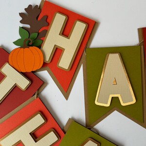 May include: A Thanksgiving banner with the word "Happy" spelled out in gold-trimmed, cream-colored letters on red and green paper flags. The letters are decorated with a textured fabric-like pattern. A small orange pumpkin and a brown leaf with green stems are attached to the "H" flag.