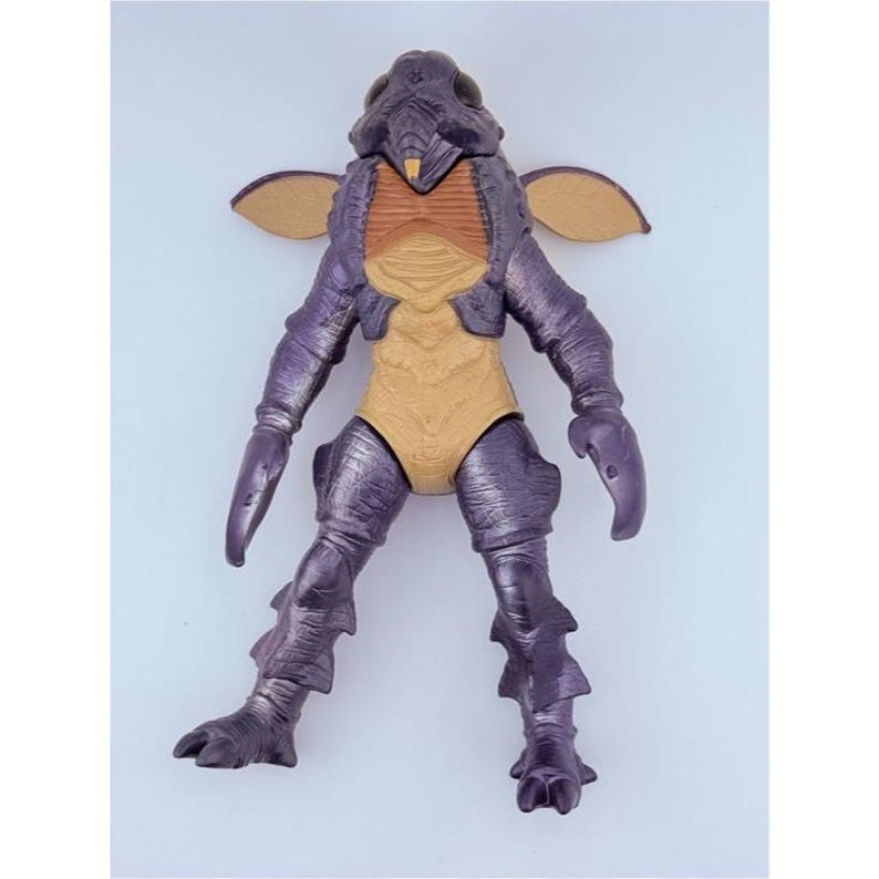May include: A purple and tan insectoid action figure with a textured exoskeleton, large dark eyes, small wing-like appendages on the shoulders, and clawed hands. The figure features a segmented body design with a tan chest and abdomen area.
