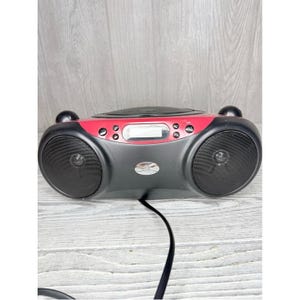 May include: A portable CD player boombox featuring a black and red plastic exterior. The device includes two circular front-facing speakers, a central digital display screen, various control buttons, and a top-loading disc compartment. A black power cord extends from the bottom.