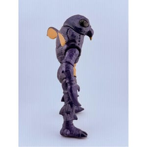 May include: A purple creature action figure with a textured, scaly body, large black eyes, and a pointed beak. The figure features tan-colored accents on the ears and chest, claw-like hands, and fin-like protrusions on the legs and back.
