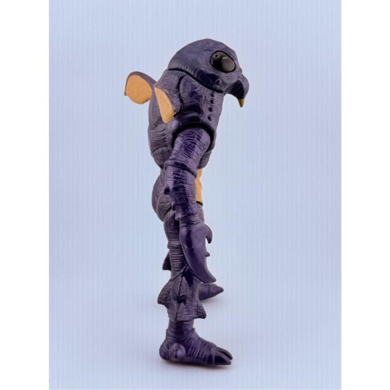 May include: A purple creature action figure with a textured, scaly body, large black eyes, and a pointed beak. The figure features tan-colored accents on the ears and chest, claw-like hands, and fin-like protrusions on the legs and back.