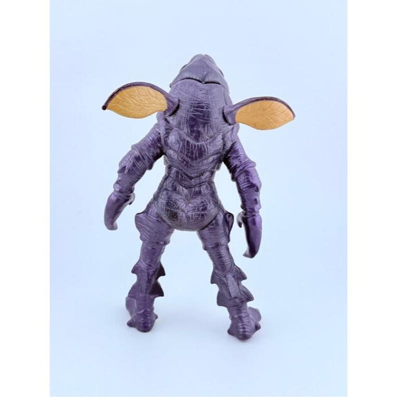 May include: A purple creature action figure with a textured, scaly body, pointed limbs, and two large, flat, tan-colored ear-like appendages extending from the sides of its head. The figure is shown from the back, displaying its detailed skin texture.