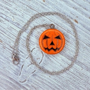 May include: A silver chain necklace with a round orange pendant carved to resemble a jack-o'-lantern. The pendant has a carved face with triangular eyes and a jagged mouth. The necklace is a Halloween-themed accessory.