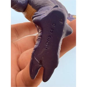 May include: A purple plastic dinosaur figurine with a textured surface. The underside of the foot is clearly marked with the text 93 BANDAI, indicating the manufacturer and year of production. The figure features a two-toned color scheme.