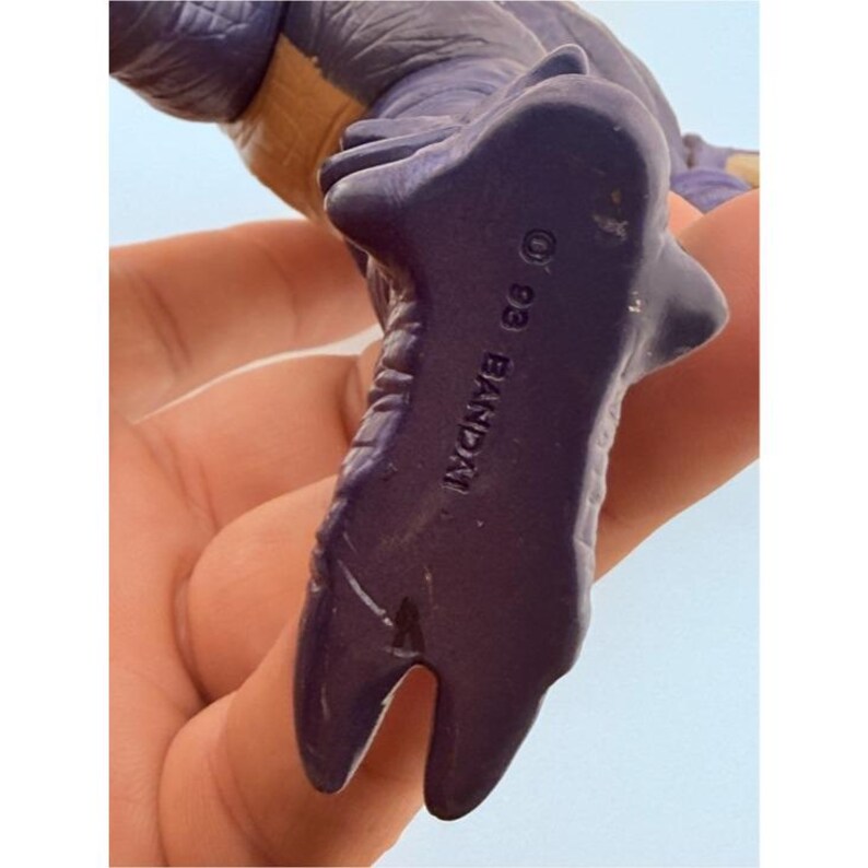 May include: A purple plastic dinosaur figurine with a textured surface. The underside of the foot is clearly marked with the text 93 BANDAI, indicating the manufacturer and year of production. The figure features a two-toned color scheme.