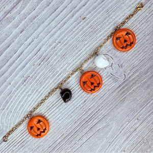 May include: A gold-colored chain bracelet featuring Halloween-themed charms. The charms include three orange jack-o'-lanterns, a white ghost, and a black bat. The bracelet is set against a light-colored wooden surface.