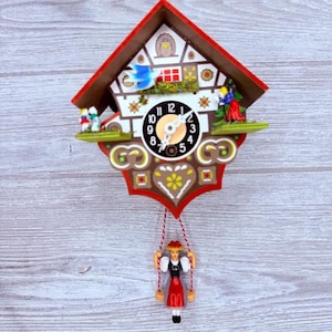 May include: A decorative cuckoo clock with a red roof and a white face. The clock features a black clock face with white numbers, a blue bird, and small figurines. A figurine swings below the clock.