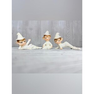 May include: Three ceramic elf figurines with pointed white hats and brown hair. One elf lies on its side, one sits upright, and one is in a crawling position. Each figure has painted facial features, including large eyes and rosy cheeks.