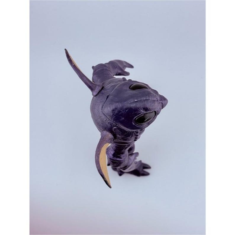 May include: A dark purple, textured creature figurine with a smooth, rounded head, large black oval eyes, and pointed, light-colored fins or appendages extending from its sides. The figure has a segmented, organic body shape and stands upright.