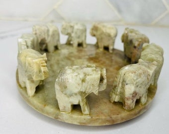 May include: A circular stone dish with a raised rim features a ring of small, carved elephant figurines. The elephants are arranged in a circle, facing inward, and are made of a light green stone.