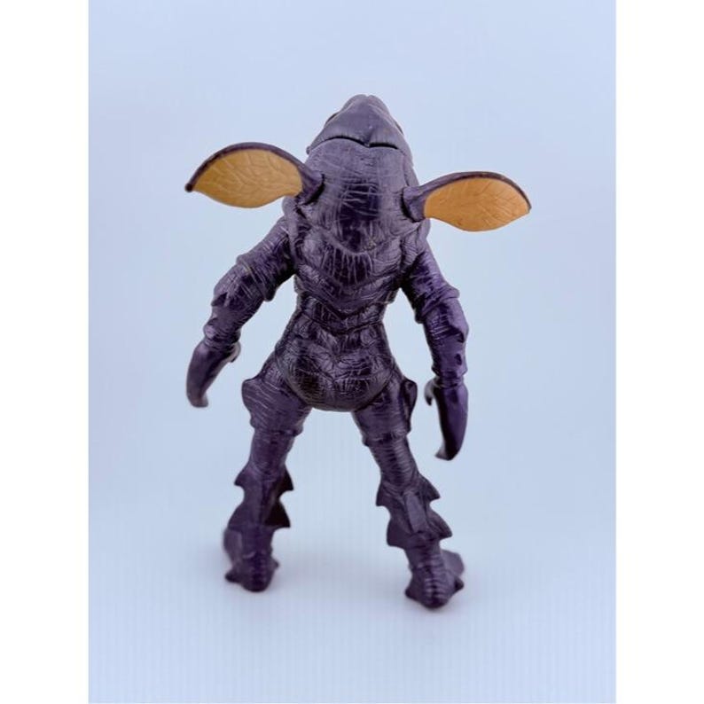 May include: A dark purple creature figurine with a textured, scaly body, pointed limbs, and two large, tan, leaf-shaped ears extending from the sides of its head. The figure stands in an upright position, showcasing a detailed, rugged surface.