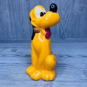 Può includere: Una statuetta di ceramica gialla di Pluto, un cane dei cartoni animati Disney. Pluto è seduto con la testa inclinata di lato e la lingua di fuori. Ha il naso nero e gli occhi neri.
