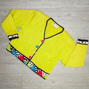 Puede incluir: Un cárdigan color chartreuse con cuello en V, ribete negro y botones coloridos. Los puños y el dobladillo presentan un patrón geométrico con acentos rojos, blancos, azules y negros. El cárdigan está hecho de un material de punto suave.