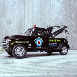 Puede incluir: Modelo de camión de remolque negro vintage con texto "Emergency Service" y "Quick Shot". El camión presenta una luz roja, un gancho de remolque y un logotipo de "Road Towing". El modelo es un artículo de colección.